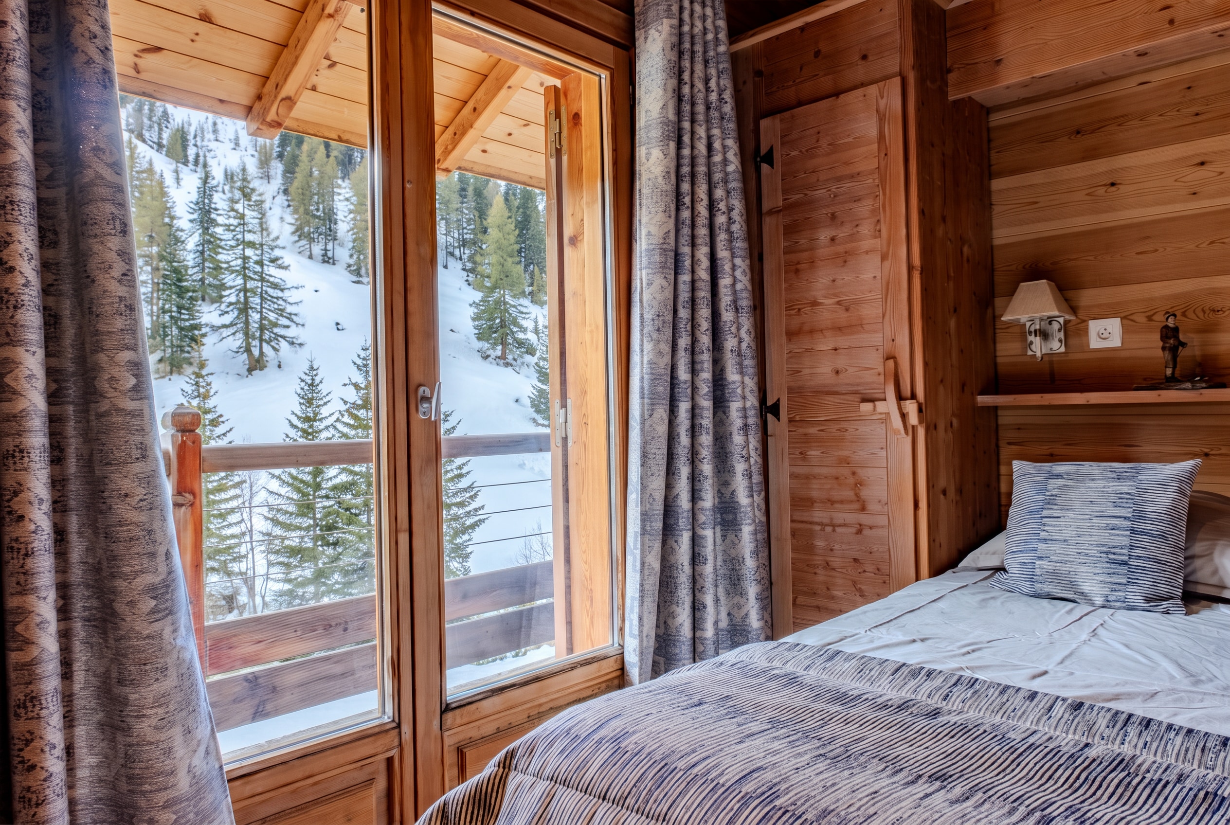 Chambre en bois avec un lit, une armoire et une porte-fenêtre ouverte sur une pente boisée