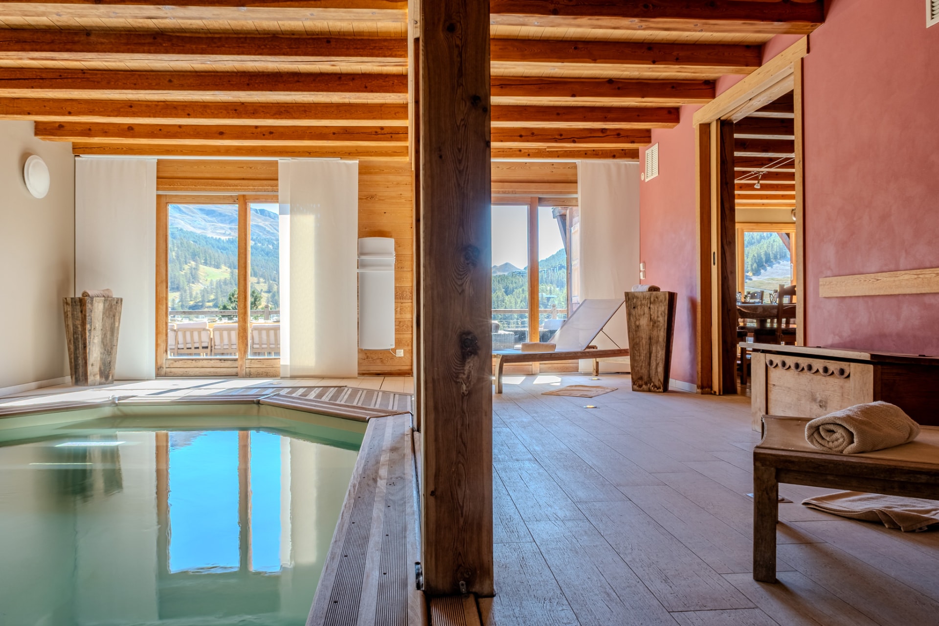 Piscine intérieure et margelle en bois, baies vitrées ouvertes sur la montagne